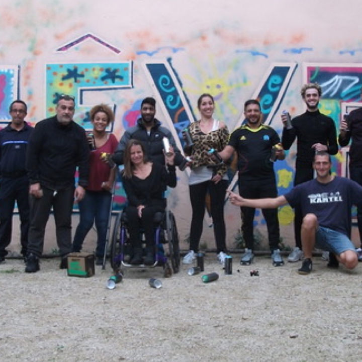 Groupe de personnes, dont une femme en fauteuil roulant, posant devant un mur décoré d’une fresque colorée avec le mot « RÊVE ». Elles tiennent des bombes de peinture et sourient, manifestant la réussite d’un atelier artistique inclusif.