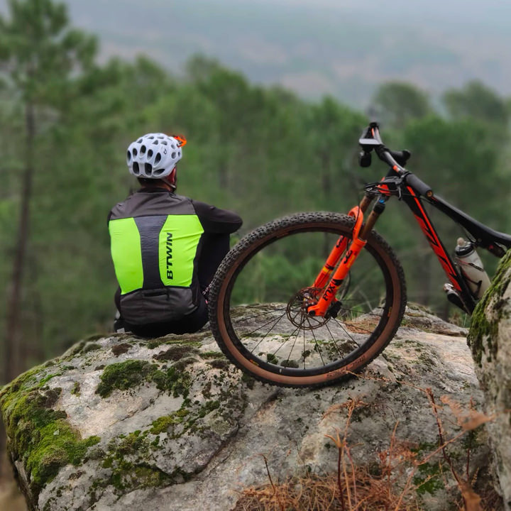 Personne portant un casque et une veste de cyclisme jaune fluo, assise sur un rocher couvert de mousse, à côté d’un vélo tout-terrain orange. Elle regarde la forêt verdoyante en contrebas, semblant profiter d’un moment de calme après l’effort.