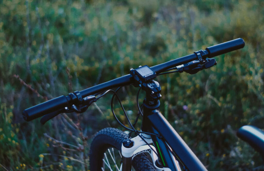 Guidon de vélo tout-terrain noir vu de près, avec compteur électronique et câbles visibles, posé dans un champ verdoyant au crépuscule.