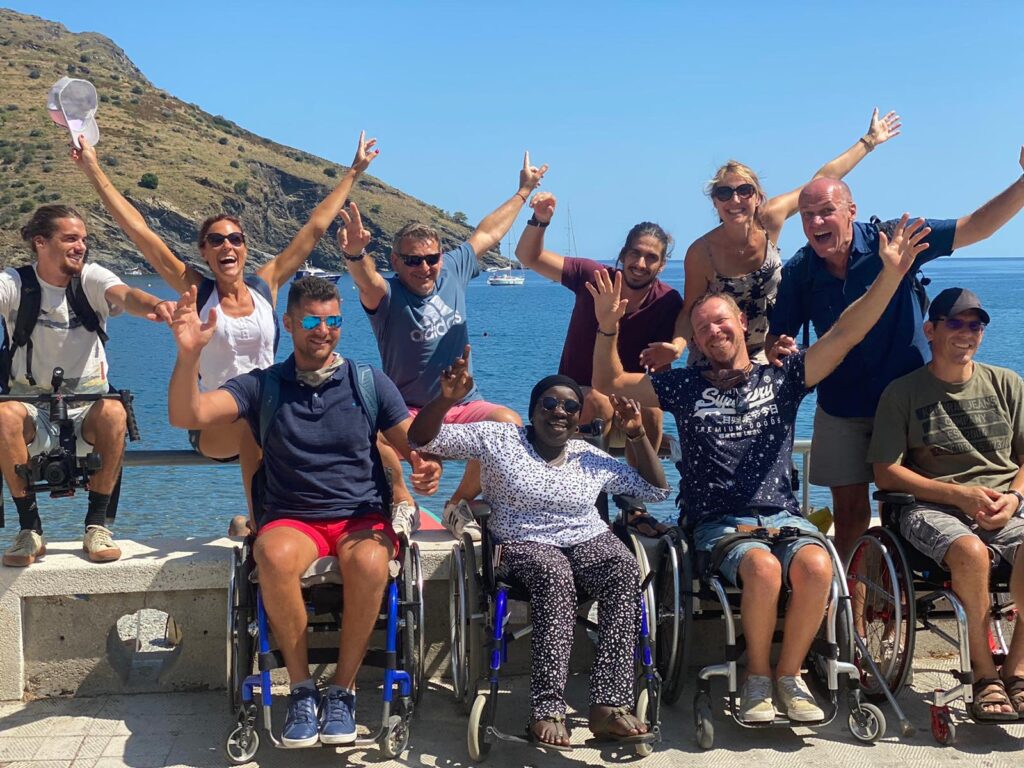 Groupe de personnes souriantes, certaines en fauteuil roulant, posant ensemble devant la mer sous un ciel bleu. Elles lèvent les bras en signe de joie, avec des collines en arrière-plan et un bateau au loin.