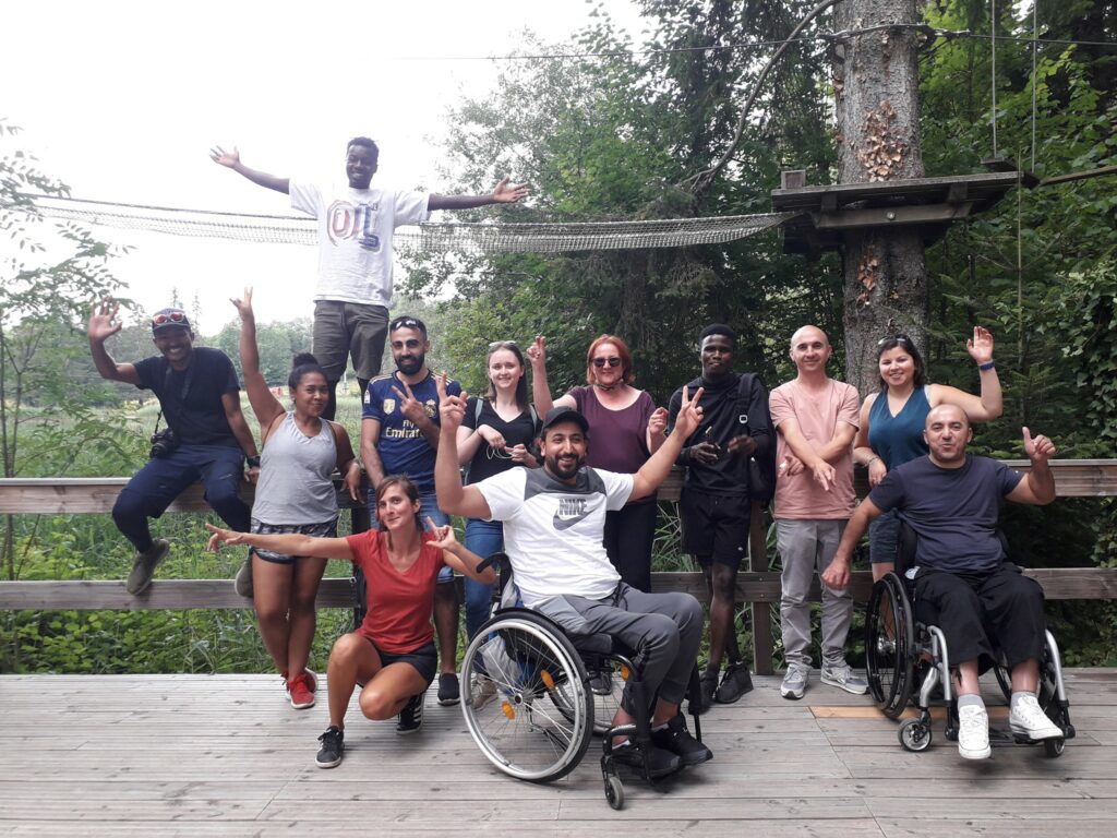 Groupe de douze personnes, dont deux en fauteuil roulant, posant ensemble sur une plateforme en bois dans une forêt. Tous sourient et lèvent les bras dans une ambiance joyeuse et conviviale, devant un équipement d’accrobranche.