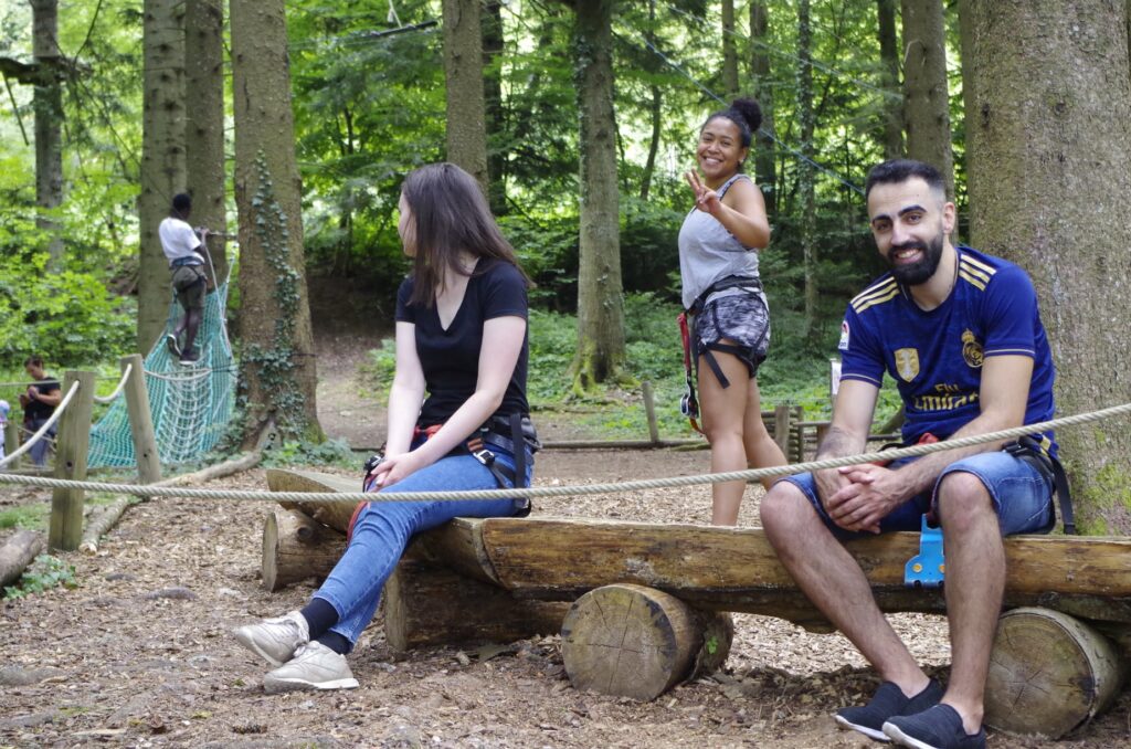 Quatre personnes participent à une activité d’accrobranche en forêt. Deux d’entre elles sont assises sur un tronc en bois, souriantes et équipées de harnais de sécurité, tandis qu’une autre salue la caméra et une quatrième grimpe sur un filet suspendu entre les arbres.