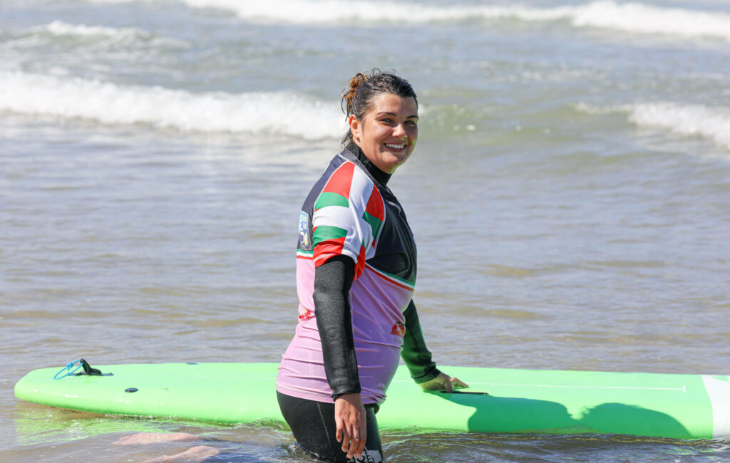 Femme souriante dans l’eau, portant une combinaison de surf et un haut coloré. Elle se tient près d’une grande planche verte, sur une plage avec des vagues en arrière-plan, par temps ensoleillé. 