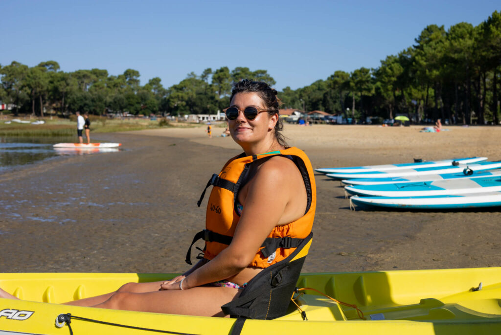 Femme assise dans un kayak jaune sur une plage, portant un gilet de sauvetage orange et des lunettes de soleil. En arrière-plan, on aperçoit des planches de paddle posées sur le sable, des personnes au bord de l’eau et une forêt de pins sous un ciel bleu.