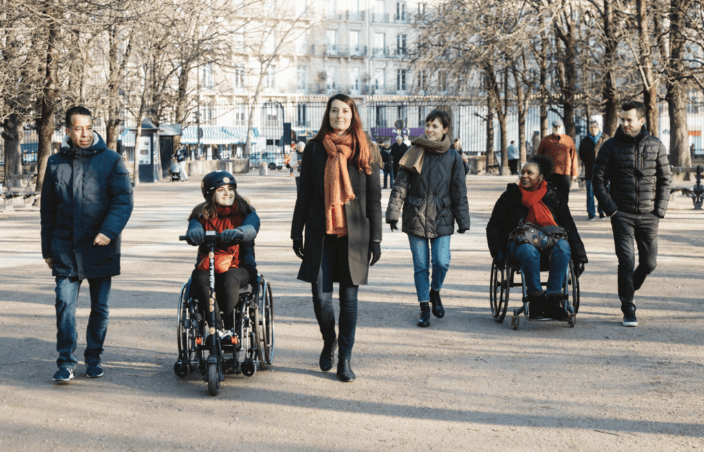 Six personnes habillées chaudement se promènent ensemble dans un parc en hiver. Deux femmes sont en fauteuil roulant, accompagnées de quatre autres personnes marchant à leurs côtés.