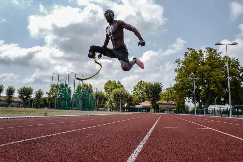 Photographie issue du film Rising Phoenix: Un athlète amputé d’une jambe effectue un saut spectaculaire sur une piste d’athlétisme, utilisant une lame de course en carbone. Il est torse nu, sous un ciel partiellement nuageux, avec des arbres et des installations sportives en arrière-plan.
