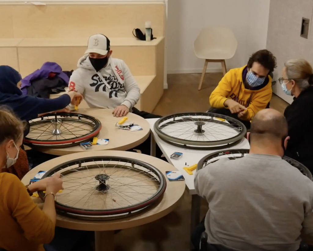 Un groupe de cinq personnes, certaines en situation de handicap, réparent ensemble des roues de fauteuil roulant autour de tables rondes dans une salle lumineuse. Tous portent des masques et échangent des conseils techniques.