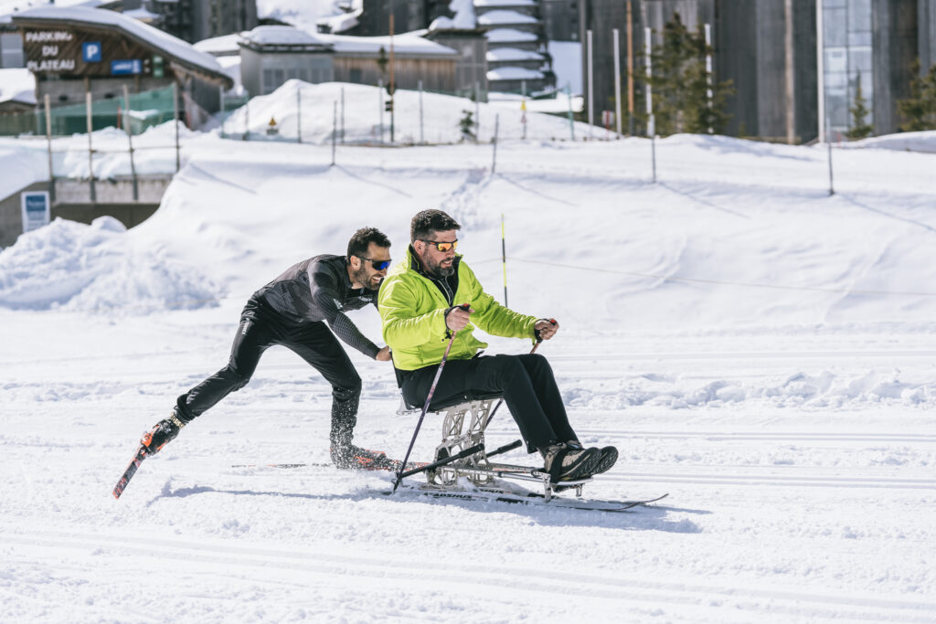 Un homme en fauteuil ski est poussé sur la neige par un autre skieur, tous deux portant des lunettes de soleil, dans une station de montagne ensoleillée.