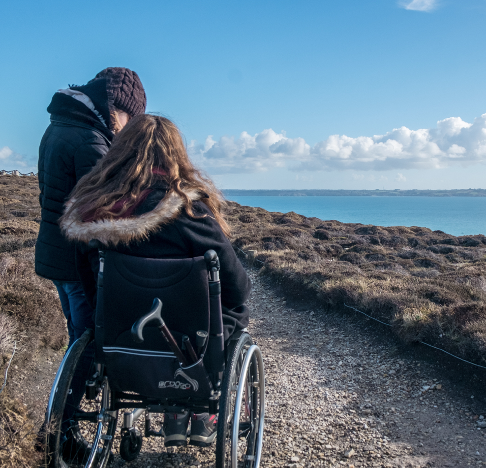 Deux personnes sont sur un sentier côtier, l’une en fauteuil roulant et l’autre debout à ses côtés, sous un ciel bleu et face à la mer. Crédit photo : www.iwheeltravel.com