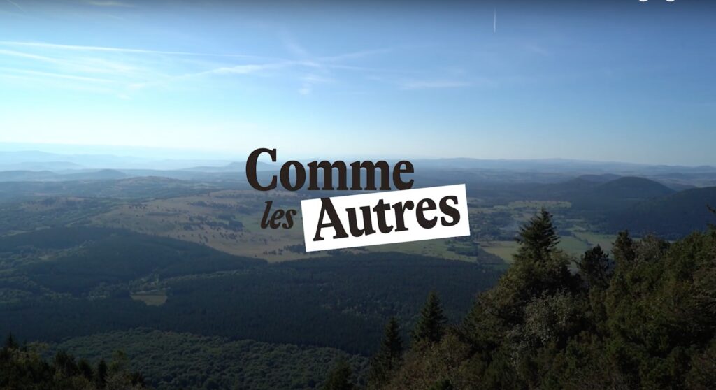 Vue panoramique d’une vallée verdoyante et de collines boisées sous un ciel bleu clair, avec le logo « Comme les Autres » au centre de l’image.