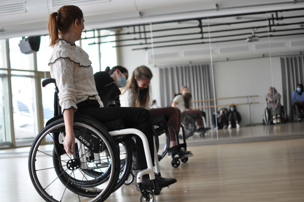 Personnes en fauteuil roulant dans une salle de cours de danse dans une salle lumineuse devant un mur de miroirs.