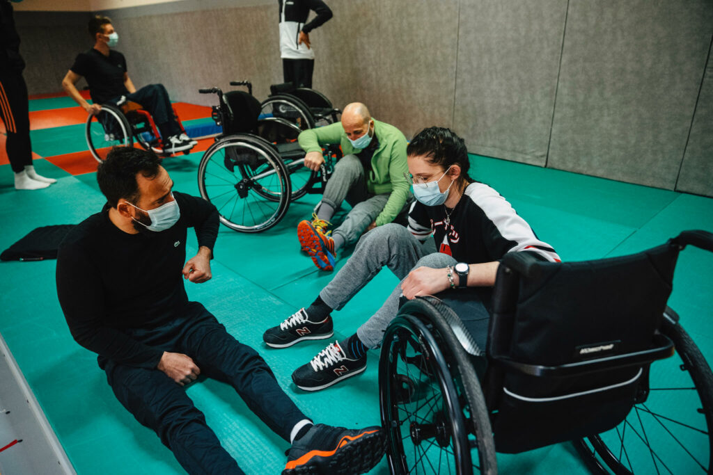 Plusieurs personnes assises au sol ou en fauteuil roulant participent à une séance de sport en salle, accompagnées d’un entraîneur. Tous portent un masque et échangent dans une ambiance conviviale.