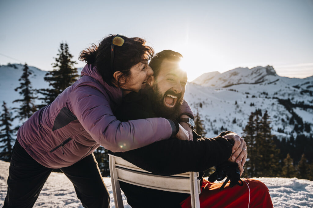 Une femme en doudoune violette enlace et embrasse un homme assis sur une chaise, tous deux riant sous le soleil des montagnes enneigées.