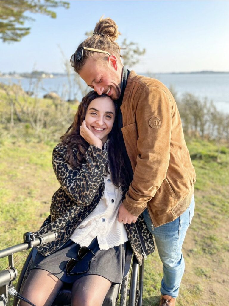 Un homme enlace une femme assise dans un fauteuil roulant, tous deux souriants, dans un paysage en bord de mer.