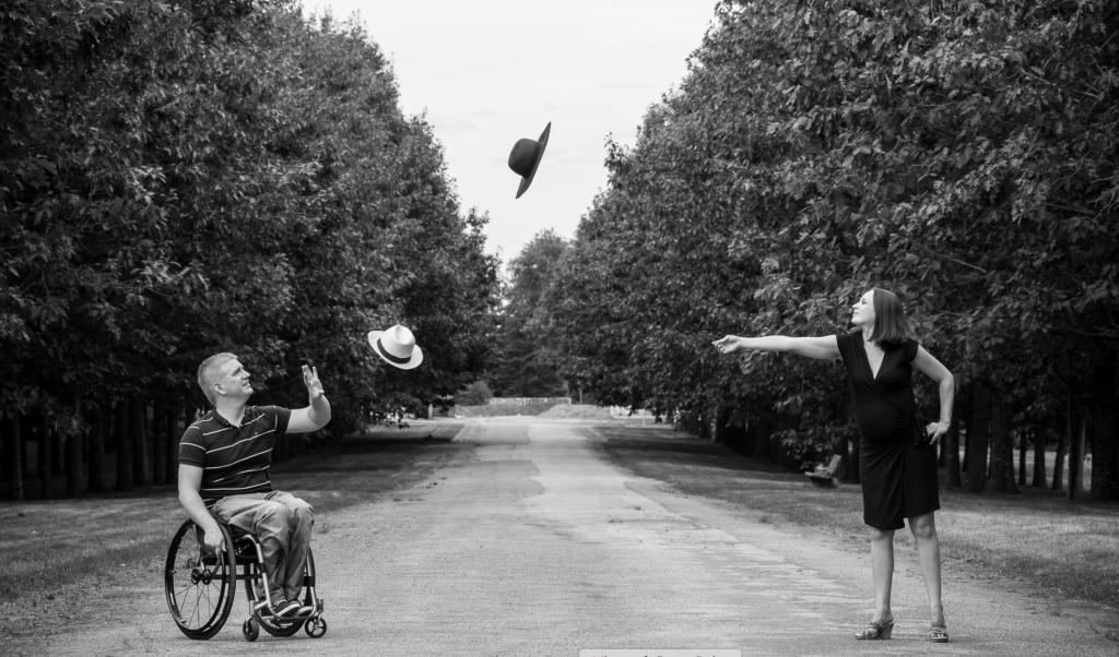 Un homme en fauteuil roulant et une femme enceinte s’amusent à lancer leurs chapeaux dans une allée bordée d’arbres.