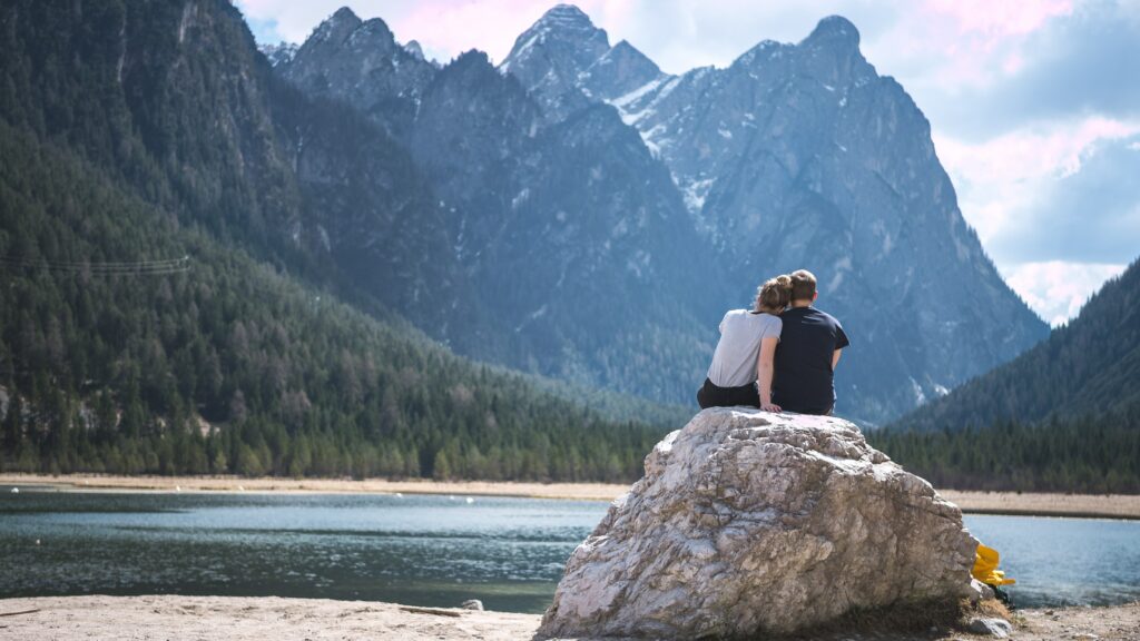 Un couple assis sur un grand rocher au bord d’un lac, face à des montagnes majestueuses sous un ciel légèrement nuageux. Photo by Timo Stern on Unsplash