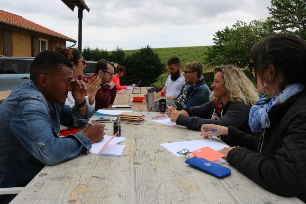 Gros plan sur une table en bois extérieure où plusieurs personnes travaillent : carnets, stylos, feuilles, gourde, téléphone et flacon de boisson à la réglisse sont disposés, suggérant une activité de groupe ou un atelier en plein air.