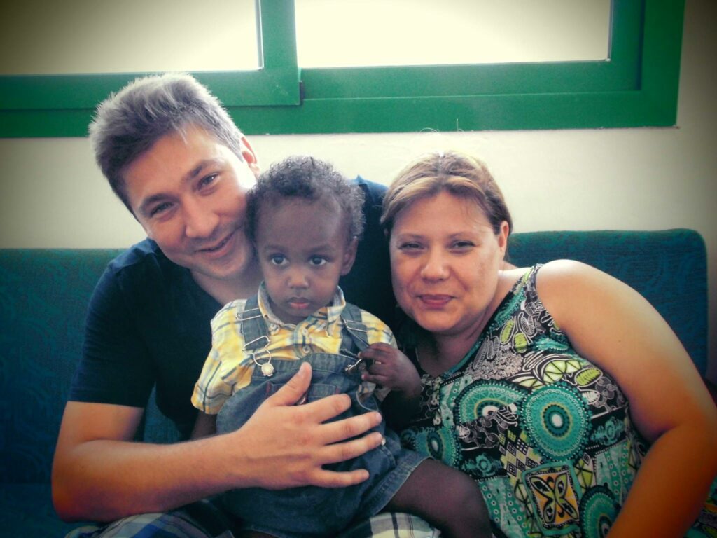 Un couple souriant assis sur un canapé tient dans ses bras un jeune enfant. L’homme, à gauche, porte un t-shirt foncé ; la femme, à droite, une robe à motifs verts et bleus. L’enfant, au centre, regarde l’objectif, vêtu d’une chemise jaune à carreaux et d’une salopette en jean. 