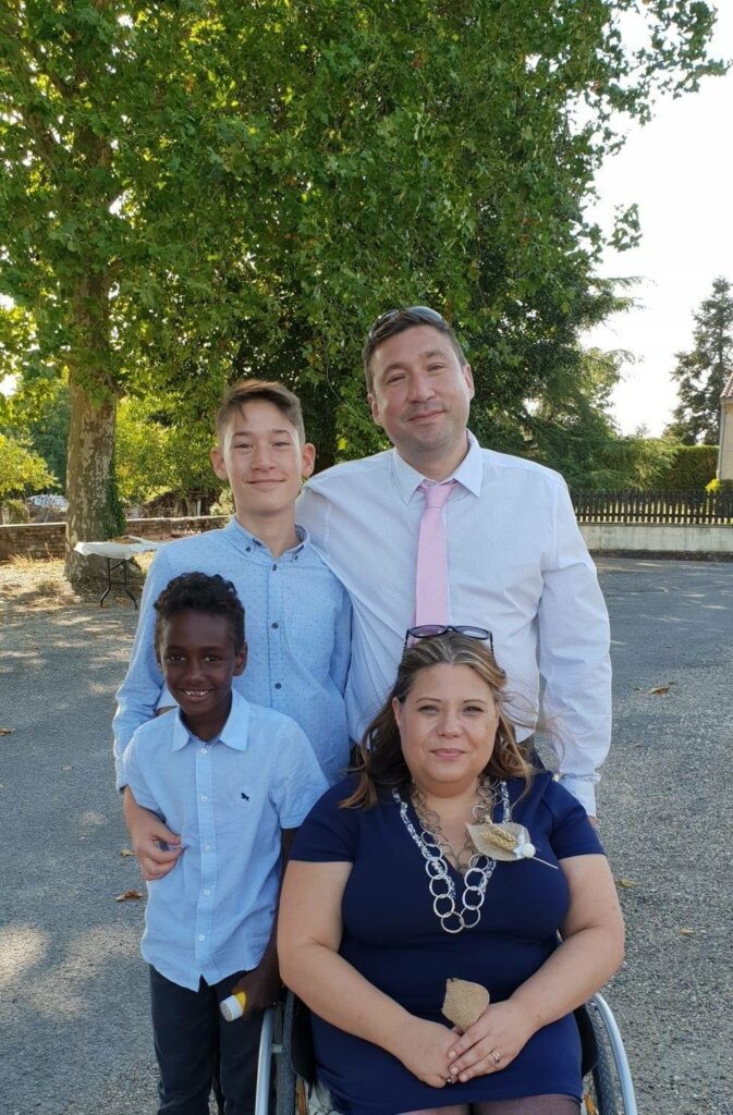 Une famille pose sur une place ombragée d'arbres: le père et les deux fils sont vêtus de chemises et se tiennent debour autour de la mère, en fauteuil roulant.