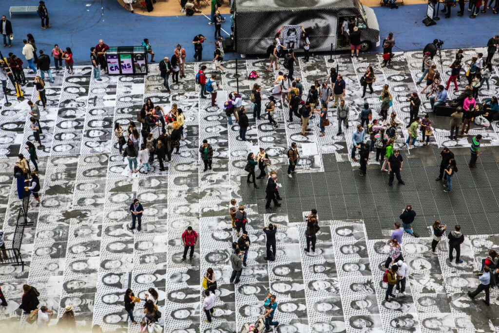 Vue aérienne d’une place recouverte de portraits en noir et blanc, où de nombreuses personnes marchent et échangent.