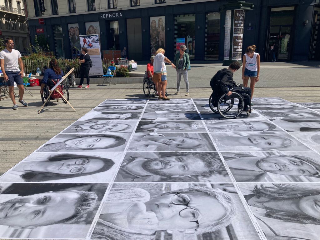 « Installation artistique en plein air sur une place urbaine : de grands portraits en noir et blanc sont disposés au sol, formant une mosaïque de visages. Autour, plusieurs personnes, dont certaines en fauteuil roulant, échangent et observent l’œuvre sous un soleil d’été, devant des boutiques