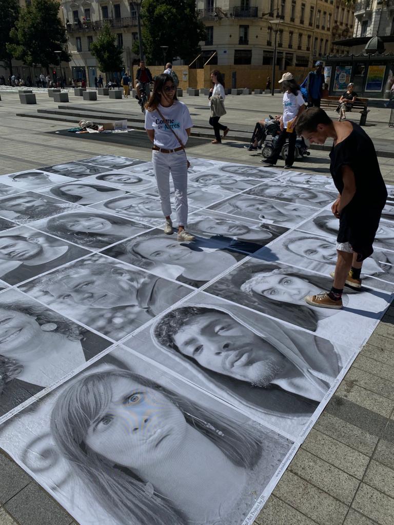 Deux personnes observent une installation artistique composée de grands portraits en noir et blanc disposés au sol sur une place ensoleillée. L’une porte un t-shirt de l’association “Comme les Autres”. En arrière-plan, quelques passants et bâtiments urbains.