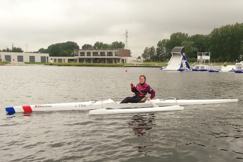 Femme souriante en tenue de sport pagayant sur un kayak blanc, sur un plan d’eau calme devant un bâtiment moderne gris et bois, sous un ciel nuageux.