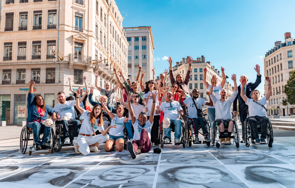 Grand groupe de personnes, dont plusieurs en fauteuil roulant, rassemblées sur une place urbaine ensoleillée. Tous lèvent les bras et sourient avec enthousiasme, portant des t-shirts de l’association “Comme les Autres”. Les immeubles historiques de la ville se dressent en arrière-plan sous un ciel bleu.