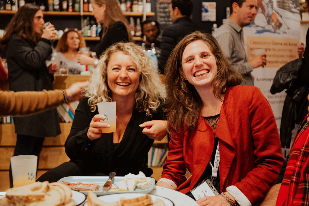Deux femmes souriantes assises à une table partagent un moment convivial lors d’un événement de l’association Comme les Autres. Devant elles, des assiettes de pain et de fromage ; en arrière-plan, plusieurs personnes discutent dans une ambiance chaleureuse. 