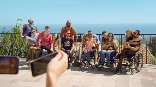 Un groupe de dix personnes, dont plusieurs en fauteuil roulant, posent ensemble sur une terrasse ensoleillée surplombant la mer. Tous sourient et s’enlacent, tandis qu’une main au premier plan tient un téléphone pour prendre la photo. 