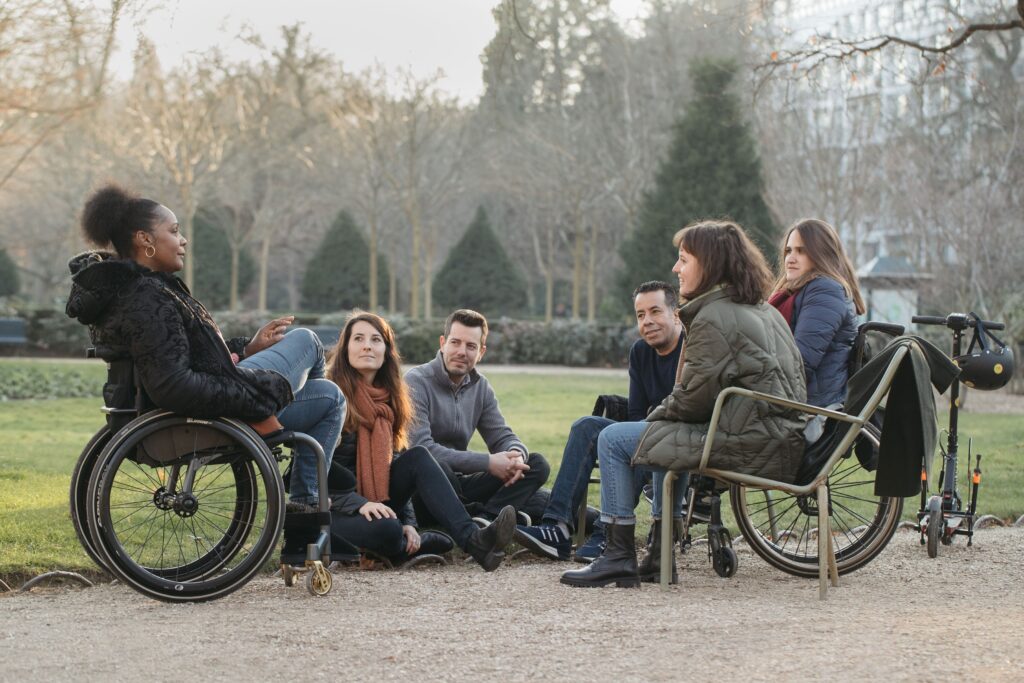 Un groupe de 6 personnes chaudement habillées, certaines en fauteuil roulant, d'autres assises par terre ou sur une chaise, discutent dans un parc.