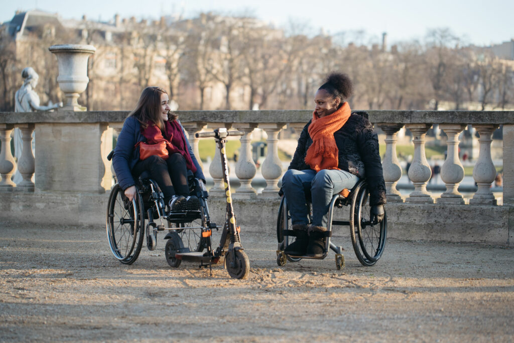 Deux femmes en fauteuil roulant discutent joyeusement dans un parc, l’une portant une écharpe orange et l’autre une veste bleue. L'un des fauteuils est couplé à une trottinette électrique. En arrière-plan, une balustrade en pierre et des arbres nus sous la lumière douce de l’hiver. 