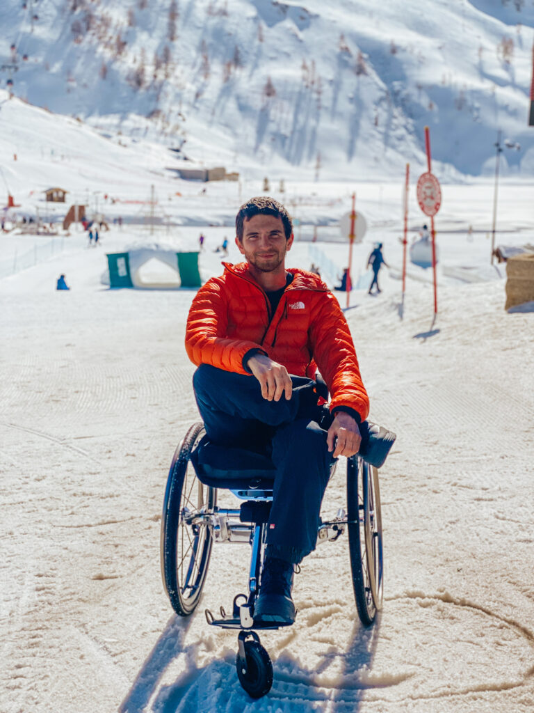 Un homme souriant en fauteuil roulant et tenue de ski rouge et noir, pose sur une piste enneigée en montagne. Derrière lui, quelques skieurs et des panneaux de signalisation se détachent sous un ciel clair.