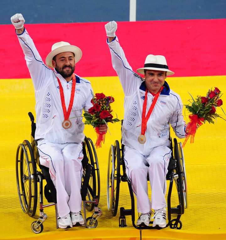 2 athlètes paralympiques portant une médaille et un bouquet de fleurs rouges saluent en levant le poing