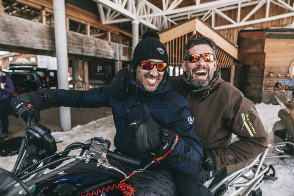 Deux hommes souriants, portant des lunettes de soleil et des vêtements d’hiver, sont assis ensemble sur une motoneige dans une station enneigée. Ils rient et posent avec complicité devant un bâtiment en bois. Crédit photo: COMME LES AUTRES © NICOLAS JACQUEMIN
