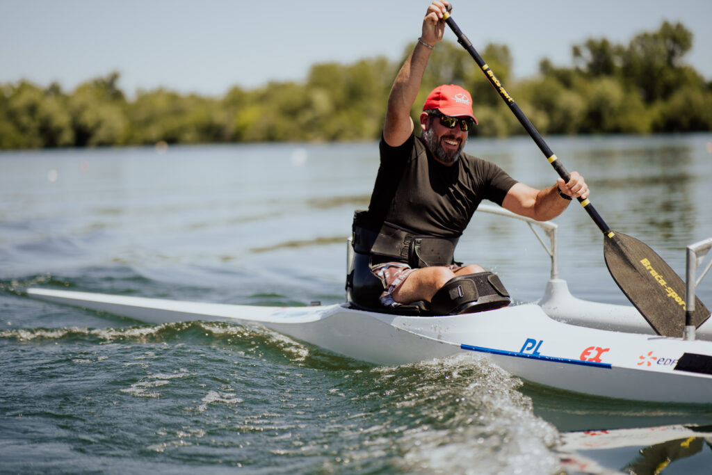 Un homme souriant pagaye sur un plan d’eau calme dans un kayak blanc équipé d’un siège spécial avec ceinture. Il porte une casquette rouge et des lunettes de soleil, sous un ciel clair entouré de verdure.