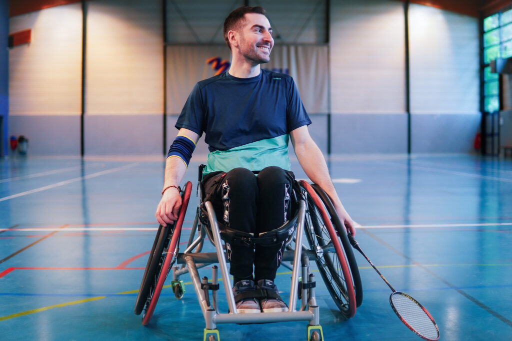 Un homme en fauteuil roulant de sport, dans un gymnase lumineux, sourit en tenant une raquette de badminton