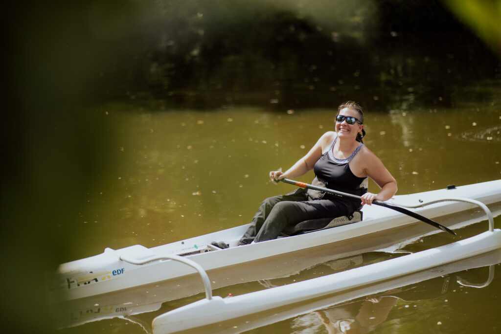 Une femme souriante rame seule sur un plan d’eau calme, assise dans un kayak blanc équipé d’un siège spécial avec ceinture, sous la lumière douce du soleil.