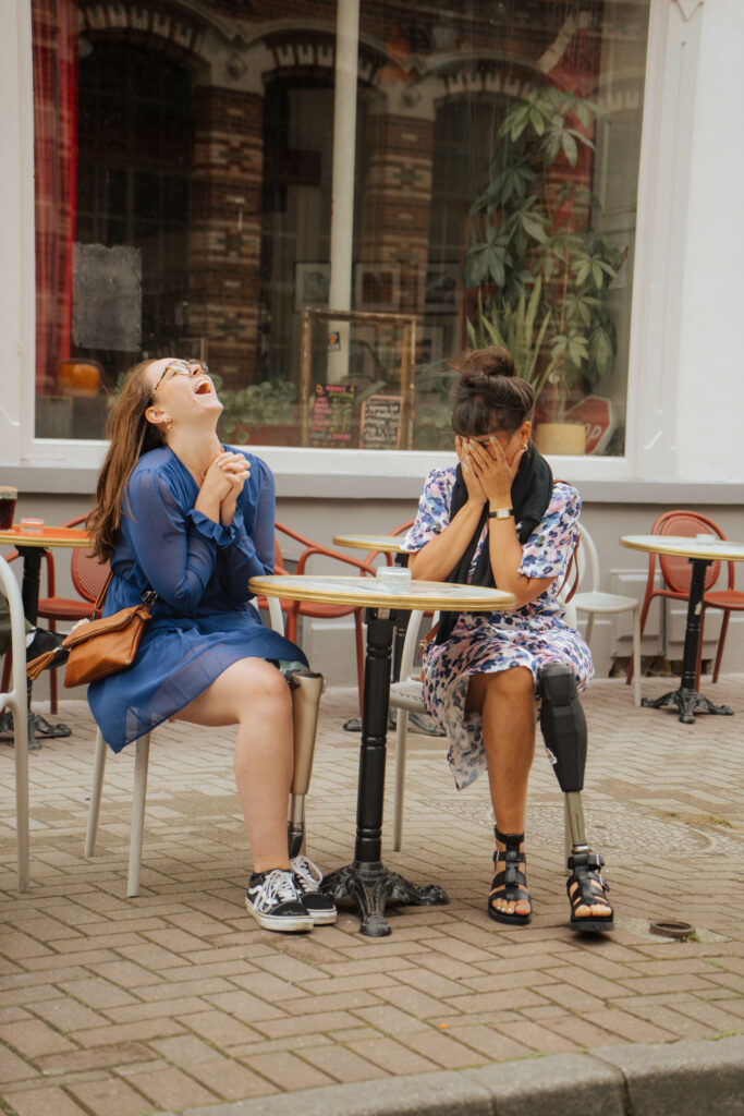 Deux femmes assises à une terrasse de café, riant ensemble. L’une porte une robe bleue, l’autre une robe fleurie et une prothèse à la jambe droite.