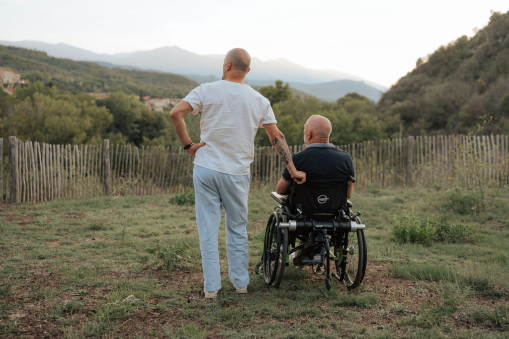 Deux hommes vus de dos, l’un debout et l’autre en fauteuil roulant, contemplent ensemble un paysage de collines verdoyantes sous une lumière douce du soir. Une clôture en bois sépare le pré de la forêt au loin.