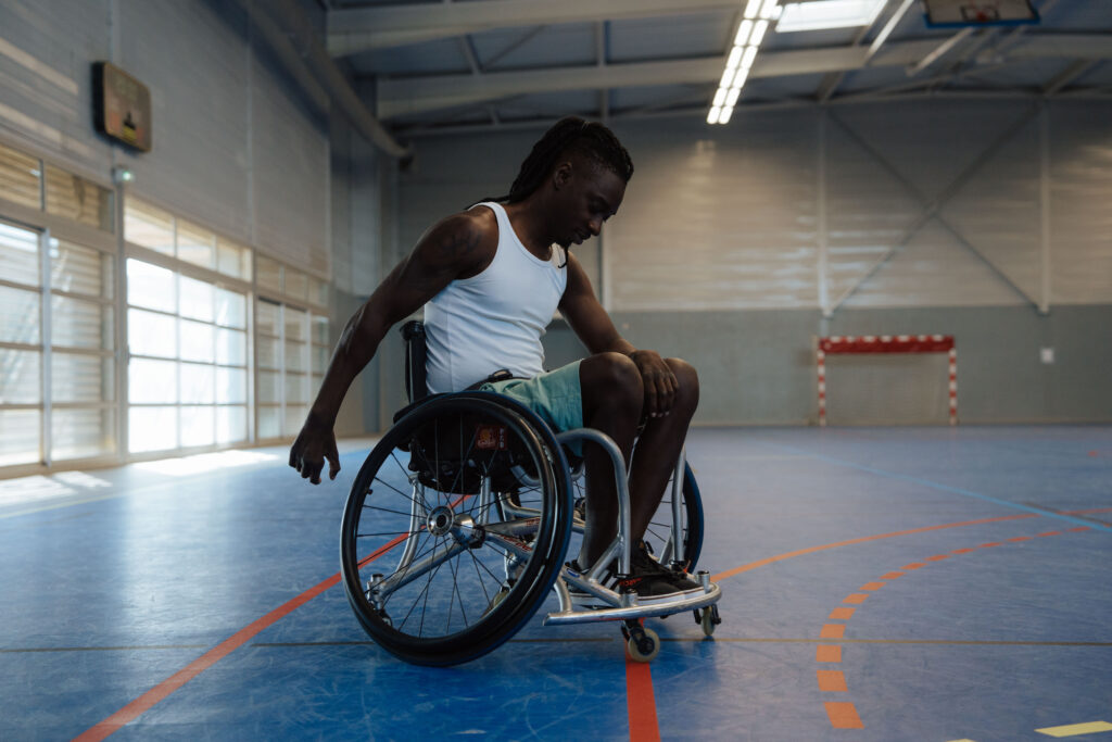 Homme en fauteuil roulant, vêtu d’un débardeur blanc et d’un short vert, s’entraînant seul dans un gymnase lumineux.