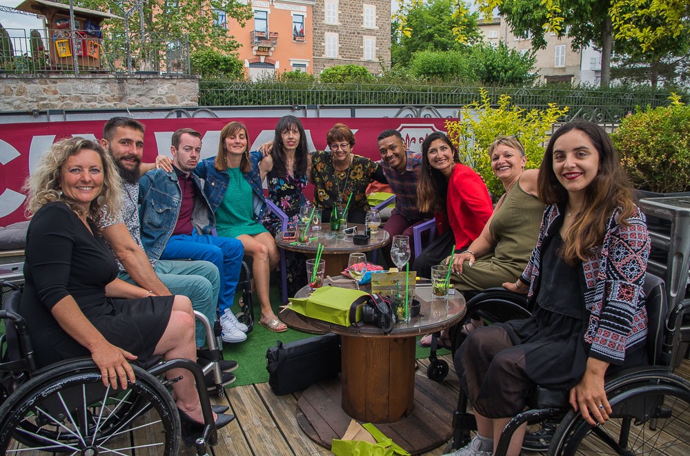 Groupe de dix personnes, dont plusieurs en fauteuil roulant, assises autour de tables en bois sur une terrasse extérieure, souriantes et partageant un moment convivial.