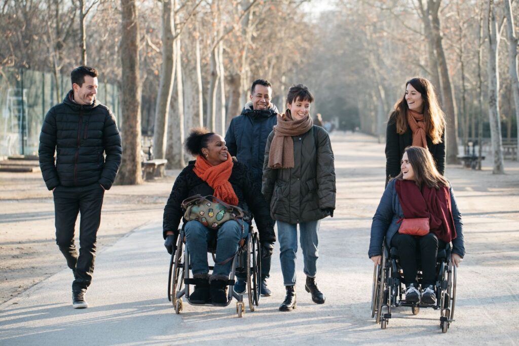 Une femme en fauteuil roulant avance sur une allée bordée d’arbres, souriante et entourée de trois amis qui marchent à ses côtés, tous vêtus de manteaux et d’écharpes par temps frais.
