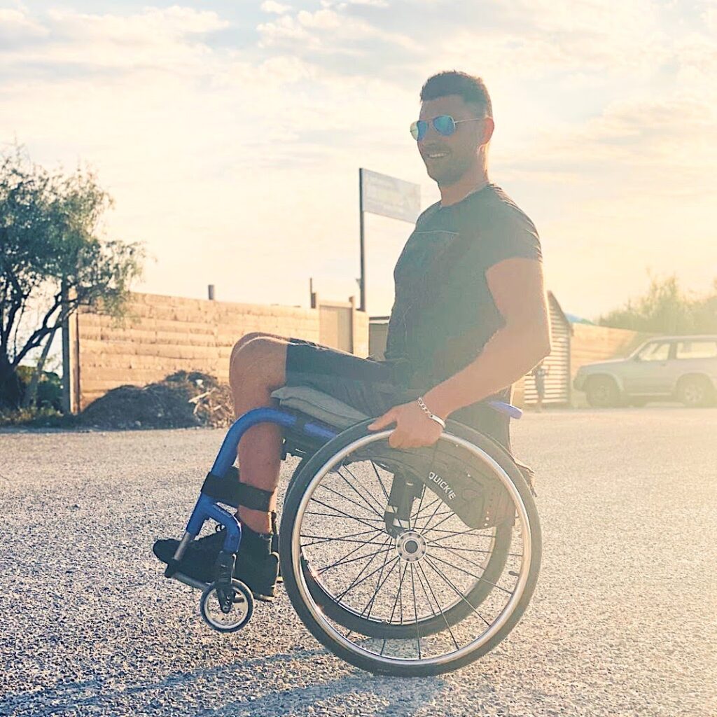 Homme souriant en fauteuil roulant sur une route ensoleillée, portant des lunettes de soleil et un t-shirt foncé, avec un fond de clôture en bois, végétation et voiture stationnée. 