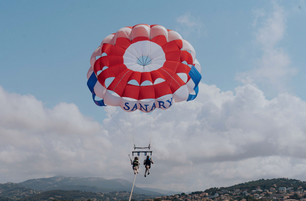 Deux personnes font du parachute ascensionnel au-dessus de la mer à Sanary, suspendues sous une grande voile rouge, blanche et bleue portant le nom “Sanary”, avec des montagnes en arrière-plan sous un ciel partiellement nuageux. 