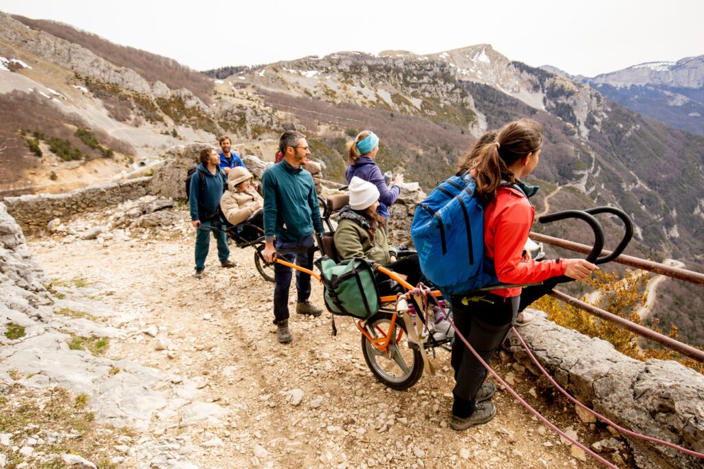  Un groupe de randonneurs aide deux personnes en situation de handicap à avancer sur un sentier de montagne escarpé, avec une vue dégagée sur les vallées et sommets environnants. 