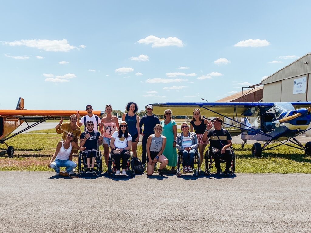 Un groupe de personnes, dont plusieurs en fauteuil roulant, posent ensemble devant deux petits avions sur un aérodrome, sous un ciel bleu et ensoleillé.