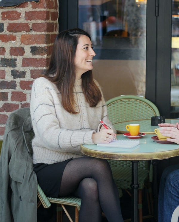 Deux femmes discutent autour d’une table de café en terrasse. L’une est assise sur une chaise, carnet et stylo à la main, l’autre est en fauteuil roulant. Toutes deux sourient et échangent dans une atmosphère détendue et conviviale.