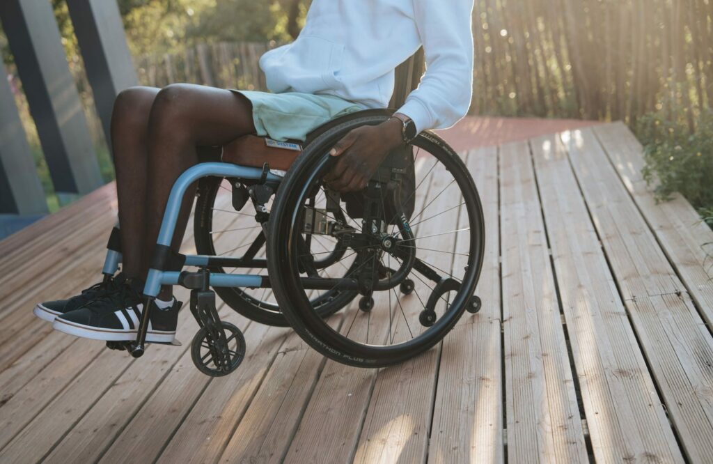Personne assise dans un fauteuil roulant sur une terrasse en bois, portant un sweat blanc et un short vert, profitant de la lumière douce du soleil.