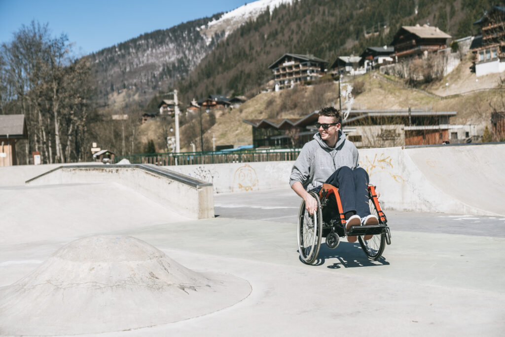 Homme en fauteuil roulant dans un skatepark de montagne, portant des lunettes de soleil et un sweat gris, souriant en se déplaçant sur la piste en béton, avec des chalets et des pentes enneigées en arrière-plan.